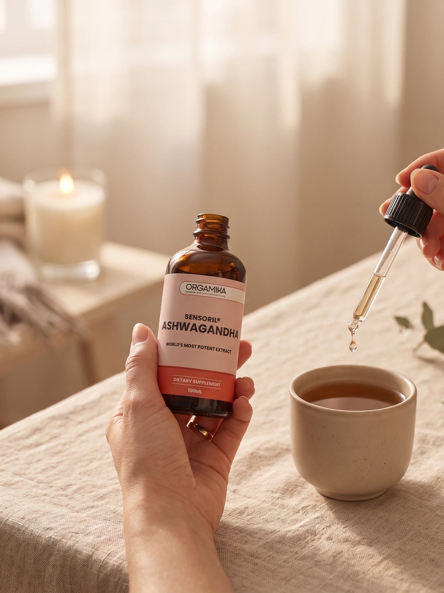 A hand holding the ORGAMIKA Liquid Sensoril Ashwagandha bottle in soft daylight, dosing a cup of tea beside a lit candle and eucalyptus
