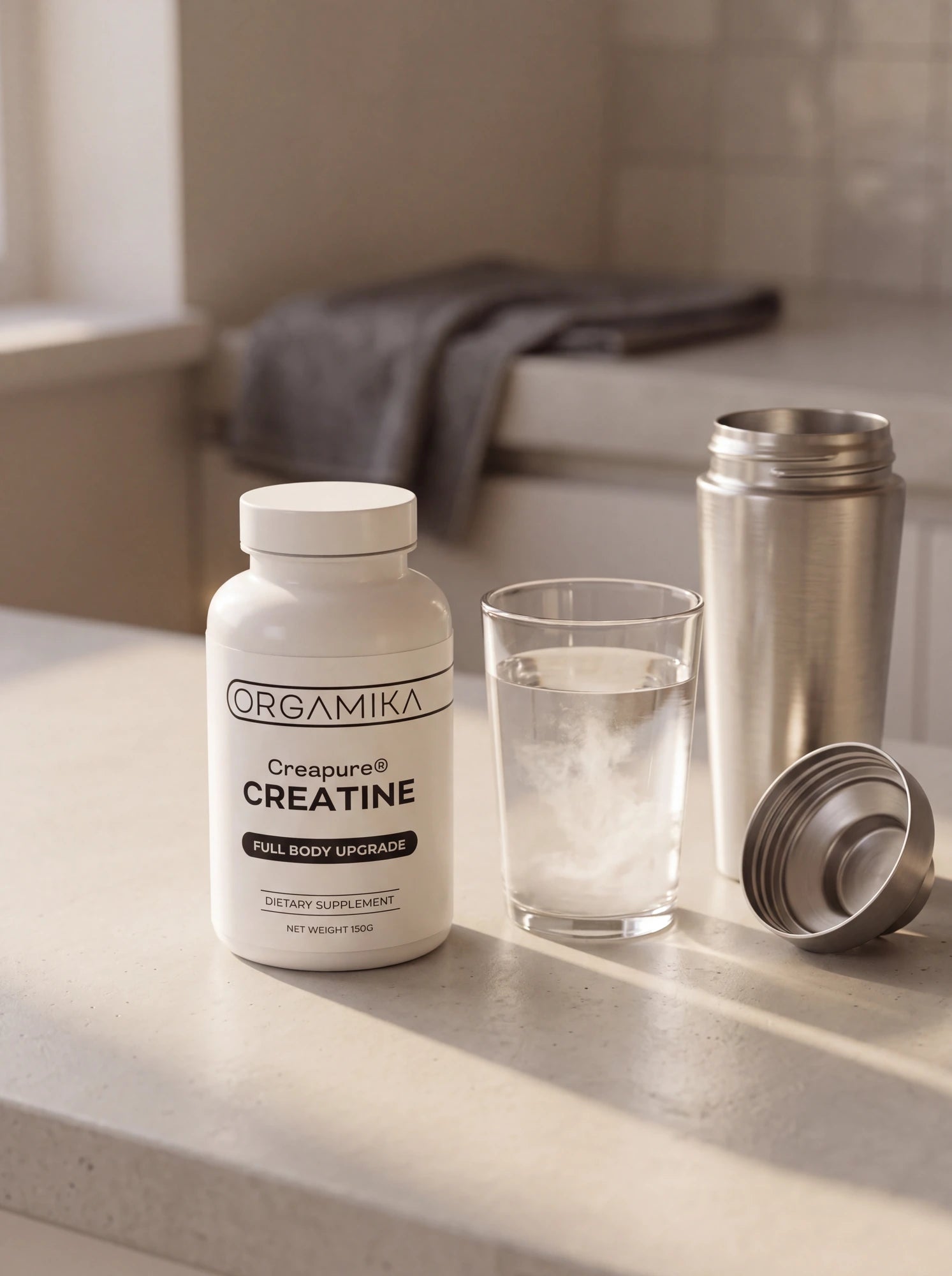 The ORGAMIKA Creapure Creatine 150g bottle on a pale stone counter beside a glass of water with creatine stirred in and a stainless steel shaker