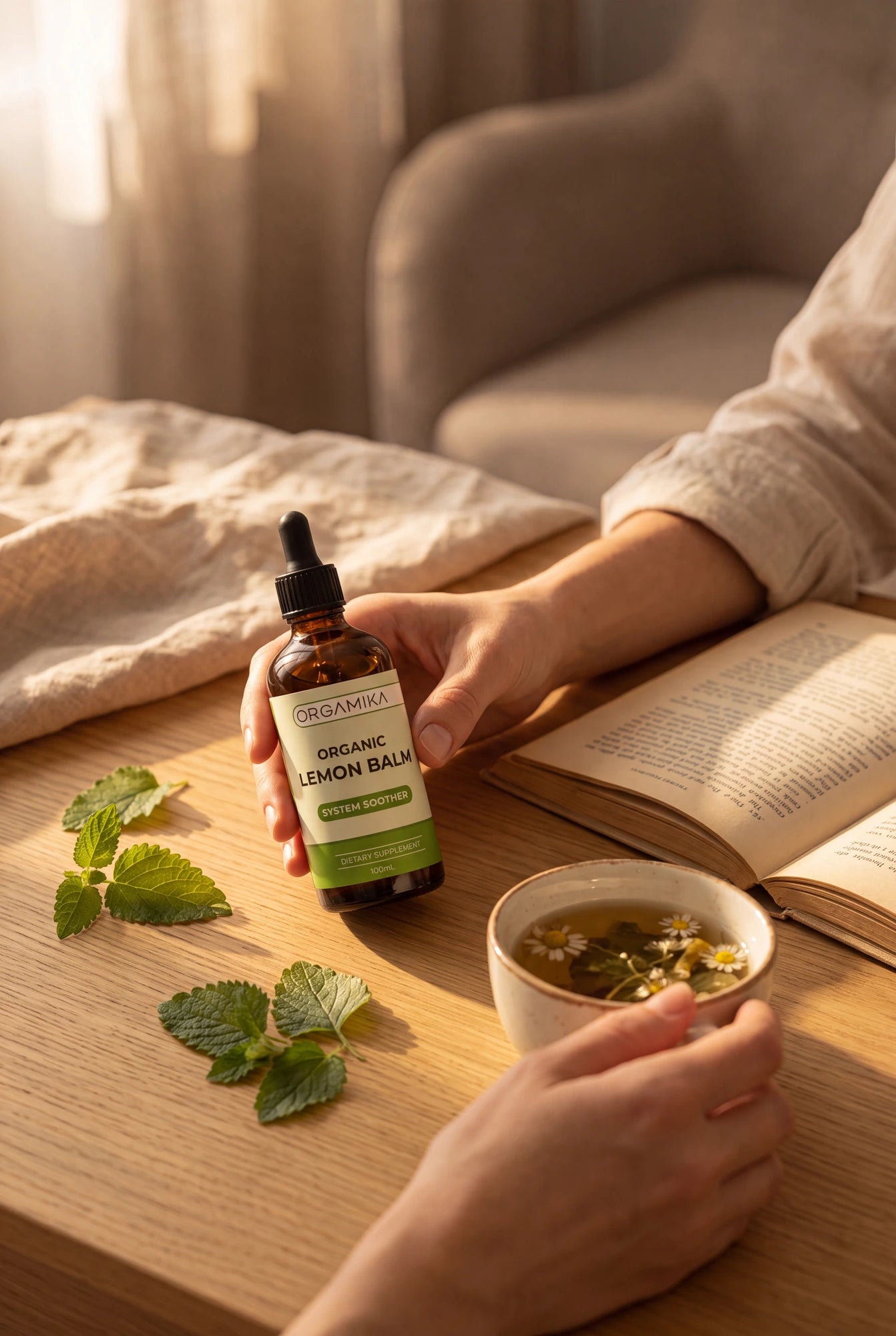 A hand holding the ORGAMIKA Organic Lemon Balm dropper bottle on a sunlit table beside an open book, a cup of herbal tea and fresh lemon balm leaves