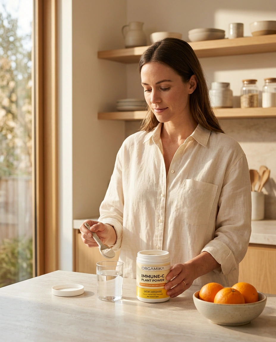 ORGAMIKA Immune-C Plant Power powder being stirred into a glass of water on a sunlit counter with rose hips and citrus alongside